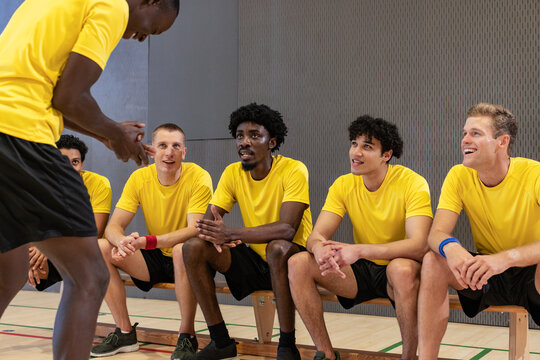 Diverse male teammates talking around wooden bench in gymnasium with court lines, yellow jerseys