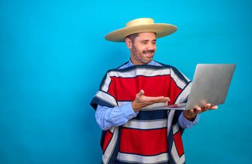Fototapeta na wymiar Happy adult latino man wearing traditional chilean huaso clothing and hat chupalla using laptop, gesturing and smiling on blue background