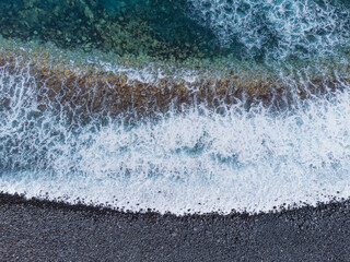 Waves crash against the volcanic shores of Tenerife, Spain, creating intricate textures in the turquoise and white waters of the Atlantic Ocean. From a drone's view