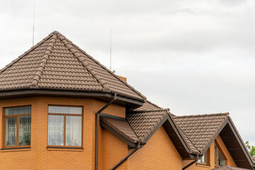 Brown tiled roof of a modern brick house with multiple peaks under a cloudy sky