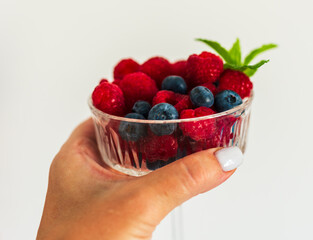 Close-up of a woman’s hand with manicured nails holding a glass cup filled with raspberries, blueberries, and mint. Natural healthy snack emphasizing freshness and well-being.