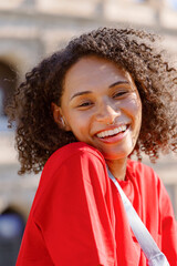 A joyful young woman with curly hair radiates happiness in a bright red shirt, smiling against a picturesque outdoor backdrop, embodying youthful exuberance and carefree enjoyment
