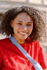A cheerful and happy young woman with beautiful curly hair, dressed in a vibrant red tshirt, smiles warmly while standing outdoors in a delightful setting against a softly blurred background