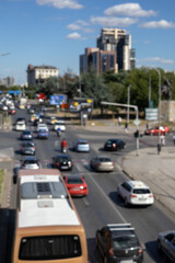 A blurry, high-angle, and vertical shot of a busy urban street with a bus and various cars. The image captures the dynamic, blurred motion of daily traffic on a sunny day in the city