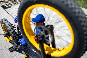 Man repairs a yellow bicycle in a sunny outdoor setting while wearing gloves and a blue cap