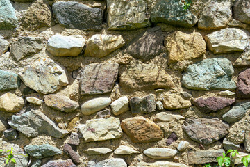 Antique wall masonry made of rough dark stones of different sizes and colors, bonded with light mortar, illuminated by sunlight