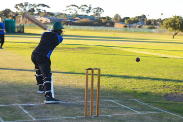 Male cricket players swinging bat at incoming ball in helmet on pitch with stumps, copy space