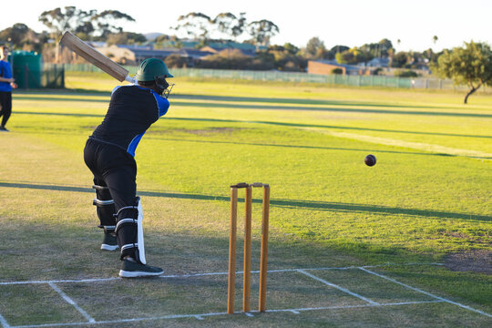 Male cricket players swinging bat at incoming ball in helmet on pitch with stumps, copy space