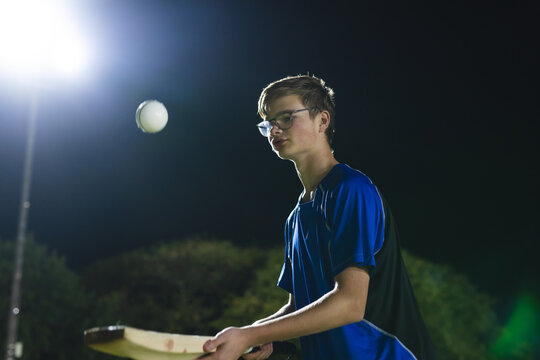 Teenage boy in blue jersey swinging cricket bat at ball on field under floodlight, copy space