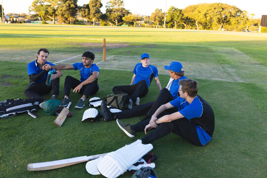 Diverse male cricket teammates sitting beside pitch in park in blue jerseys with helmets