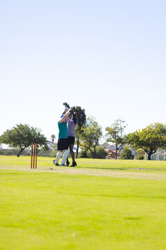 Diverse male teammates in pads and gloves giving high five on cricket pitch near wooden stumps