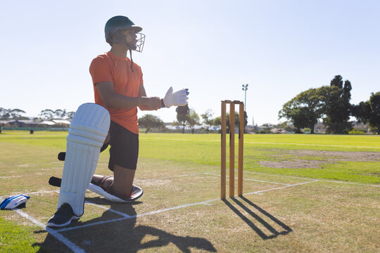 Cricketer in green helmet kneeling grass pitch adjusting glove strap by wooden stumps