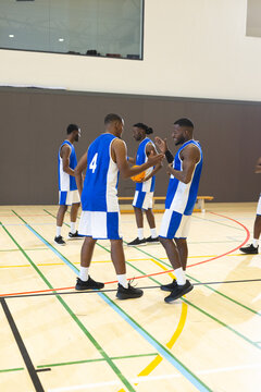 African american male teammates passing basketball, exchanging handshake on gym court by bench