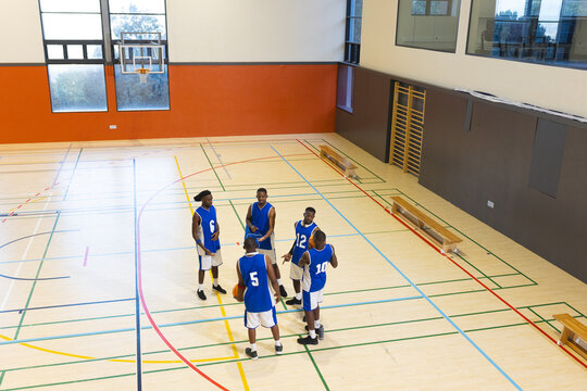 African american male teammates discussing basketball play on gym court next to benches and hoop
