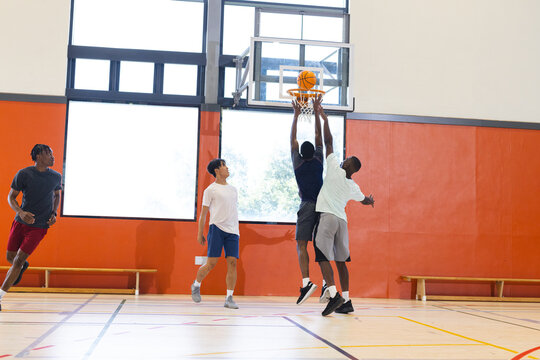 Diverse male friends jumping for basketball under hoop at gym on polished court with benches - Powered by Adobe