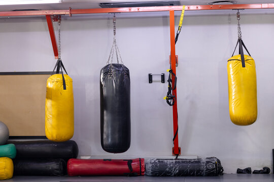 Three punching bags hanging from red beam on gym gray wall beside mats, trx straps