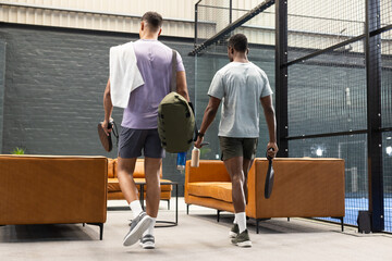 Diverse male friends walking toward paddle court from lounge carrying rackets duffel water