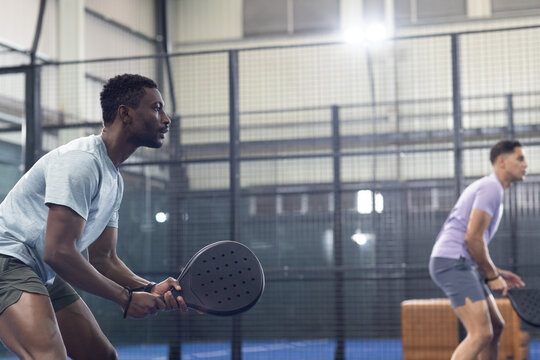 Diverse male teammates standing on paddle tennis court with rackets next to mesh walls, copy spac - Powered by Adobe