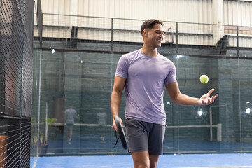 African american man in lavender shirt holding padel racket tossing tennis ball on padel court