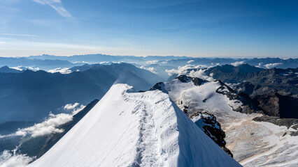 Hikers navigate the steep and sharp ridge of Piz Palu, surrounded by breathtaking views of the Swiss Alps, clouds over valleys, and glaciated terrain during summer ascents.