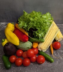 On the gray table there are fresh juicy vegetables: zucchini, avocado, tomatoes, cucumbers, bell peppers, corn, lettuce leaves