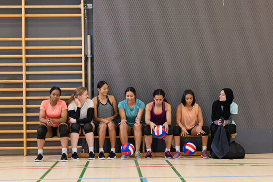 Diverse female volleyball teammates sitting on wooden bench in gym with tricolor volleyballs