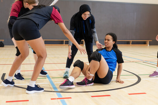 Diverse female volleyball team helping injured teammate in knee pads holding ankle on court - Powered by Adobe