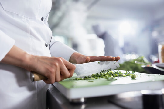Focused woman chef slicing fresh herbs for a new gourmet dish on the stove, cutting rosemary or basil at the kitchen stove. Female expert using raw materials and ingredients for a meal.