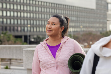 Woman standing outside office building on plaza holding green yoga mat, wearing pink jacket