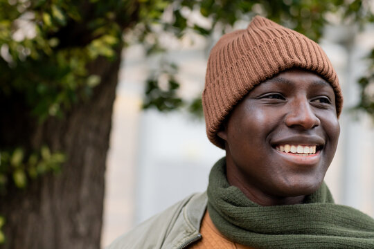 African american man standing by tree trunk wearing jacket, beanie and scarf on walkway