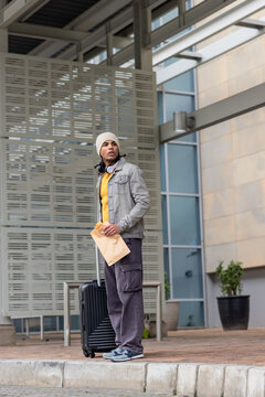 Asian man waiting outside stone-clad building entrance holding suitcase and paper bag