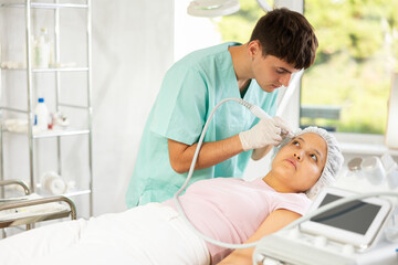 Obraz premium Young male cosmetologist performs hardware facial cleansing procedure to young woman patient