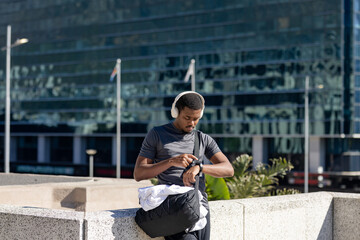 African american man in 20s leaning against wall, gym bag in city plaza checking smartwatch
