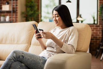 Obraz premium Cheerful female entrepreneur sitting on couch in modern apartment, using smartphone and debit card for digital payment. Asian businesswoman completing online purchase on mobile device.