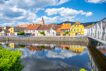 Obraz premium Vibrant buildings line the Otava River in Susice, showcasing bright colors and clear reflections on the water. A bridge connects both sides under a blue sky filled with clouds.