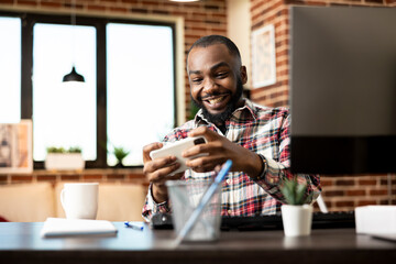 Cheerful black male freelancer having fun with mobile gaming at home office desk. African american remote manager holding cellphone with excited expression and playing video games during break time.