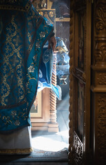 Priest in beautiful blue vestment at Divine Liturgy on Feast of the Virgin in Orthodox church, censer and incense smoke in sunlight at altar