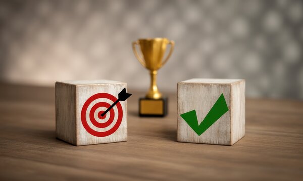 Two wooden blocks, one with a target and arrow, the other with a checkmark, sit on a wooden surface near a golden trophy