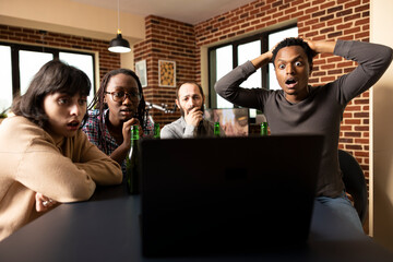 Shocked black man with hands on head, watching online content with friend, gathered around table in brick wall room. Young diverse adults reacting with wide eyes to suspenseful scene on laptop screen.