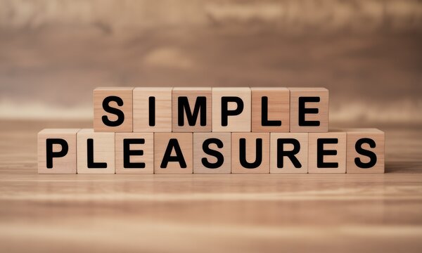 Wooden blocks spelling out "SIMPLE PLEASURES" on a light brown wooden surface