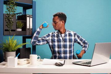 Black male entrepreneur training his muscles by lifting dumbbells and using laptop at office desk. African american man multitasking fitness and business, staying active during his working hours.