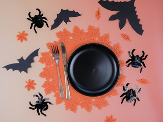 Top view empty black plate on round orange felt placemat with knife fork autumn leaves and Halloween decorations on peach background