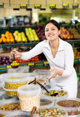 Young woman shopper in casual clothes putting olives in plastic packaging in supermarket..