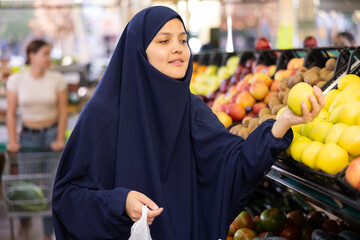 In vegetable shop,middle-aged woman in traditional Muslim hijab takes and puts ripe apples in small bag.Woman is shopping in background