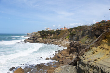 Rocky coastline and ocean waves at Praia das Ma&ccedil;&atilde;s, Portugal. Scenic view of the Atlantic coast with cliffs, beach, and seaside houses under a bright sky. Perfect image for travel, tourism