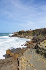 Rocky coastline and ocean waves at Praia das Maçãs, Portugal. Scenic view of the Atlantic coast with cliffs, beach, and seaside houses under a bright sky. Perfect image for travel, tourism