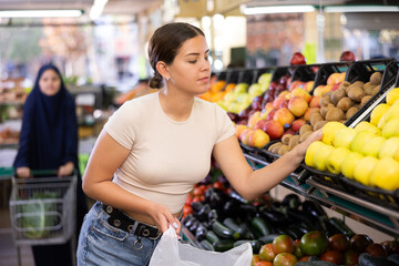 Young woman shopper in casual clothes chooses apples in supermarket