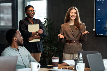 Business Presentation. Smiling Young Businesswoman Giving Speech During Seminar With Coworkers In Office, Standing At Desk In Boardroom, Diverse People Sitting At Table And Listening To Happy Speaker