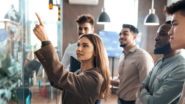 Portrait of young businesswoman mentor coach leader writing idea or task pointing at sticky notes on glass wall, diverse team developing work plan in creative corporate office at stand up meeting