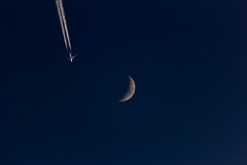 A plane in the night sky with the moon and contrail.